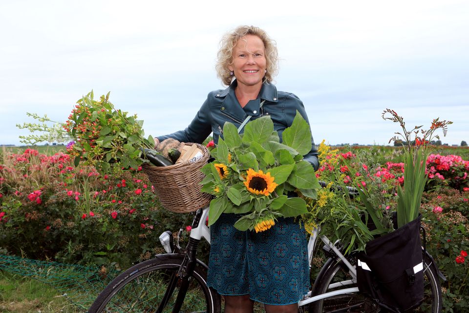 Een mevrouw met blonde krullen staat bij haar fiets in een bloemenveld en ook in het fietsmandje zitten bloemen.