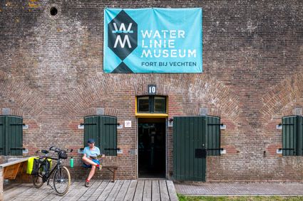 Een man zit op een bankje voor een bakstenen fort met grone luiken voor de ramen en deuren. Naast hem staat zijn sportfiets. Boven de deur van het fort hangt een blauwe banier met daarop: Waterliniemuseum Fort bij Vechten.