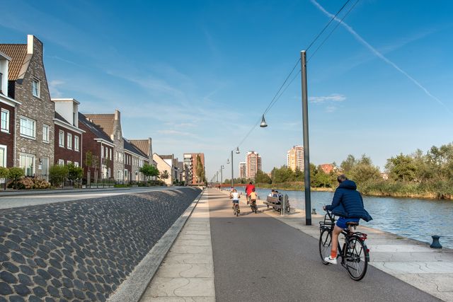 Foto van het wandel- fietspad langs het water in Oosterheem. Er fietsen wat ouders en kinderen langs het water.