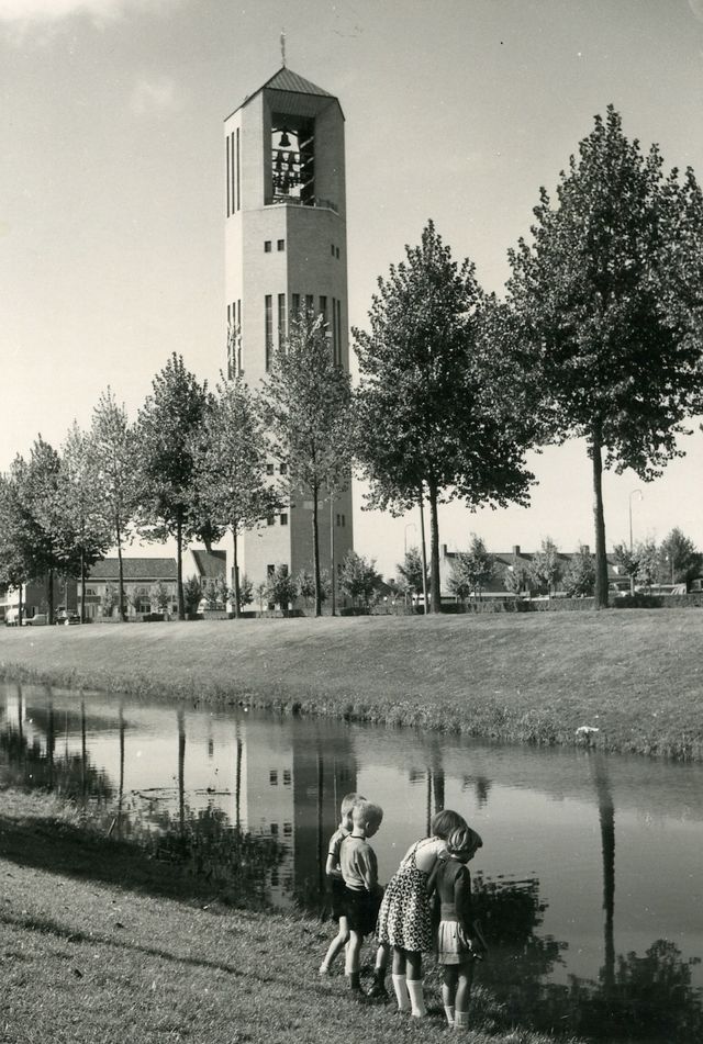 Zwart wit foto van een toren in Emmeloord in de Noordoostpolder waar een paar bomen omheen staan. Op de voorgrond zie je enkele kinderen bij het water staan kijken.