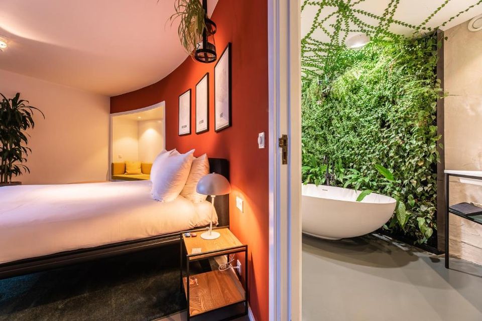 Suite in Hotel Gooiland met oranje wand achter het bed, en doorkijkje naar de badkamer met vrijstaand bad en wand vol planten.