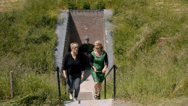 Twee vrouwen komen de bakstenen gang van een fort uitgelopen naar buiten. Achter de vrouwen zie je hoe het fort bekleed is met aarde en overgroeid met gras.