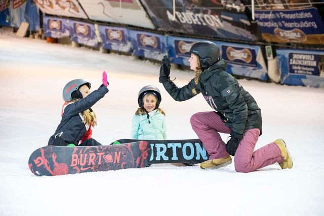 Twee kinderen liggen met hun snowboard en geven de instructeur die ook een snowboard aan heeft een high five.