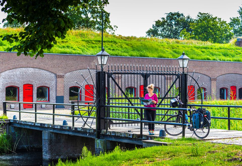 Een vrouw staat met haar fiets op de brug voor de poort van een fort van baksteen met rode luiken. Ze bekijkt een folder.