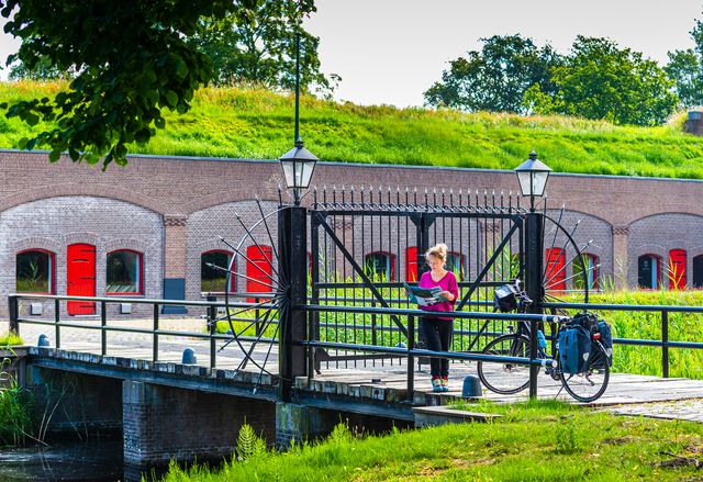 Een vrouw met een fiets staat buiten de poort van een bakstenen fort met rode luikjes. Ze kijkt op een kaart.