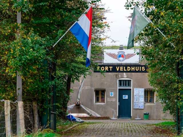 Een betonnen fort met daarvoor bossages. In de bomen hangen twee vlaggen. Op het fort staat geschreven: Fort Veldhuis.