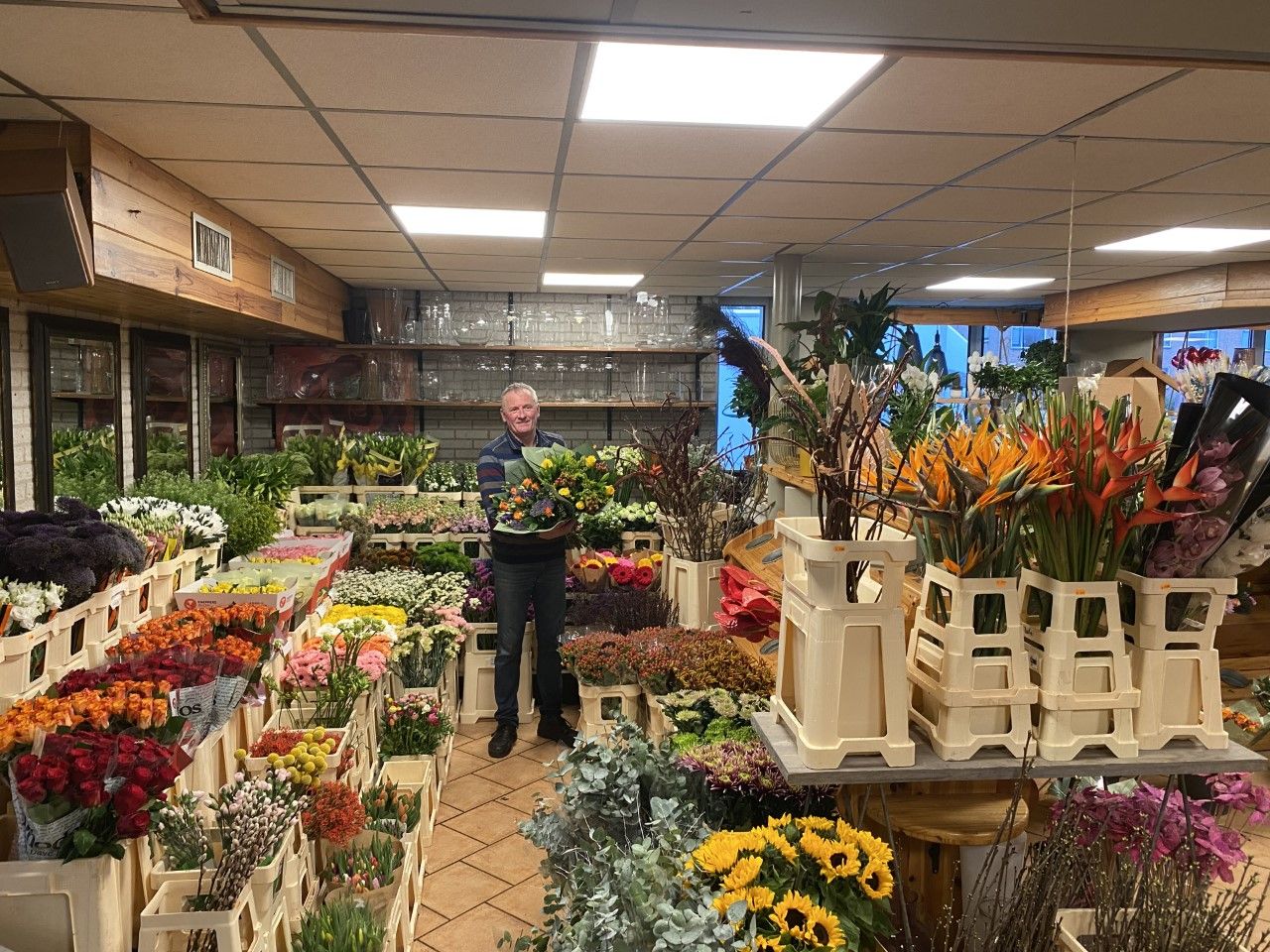 Een man met een bos bloemen in de hand terwijl hij in een bloemenzaak staat met allemaal fusten met verschillende bloemen.