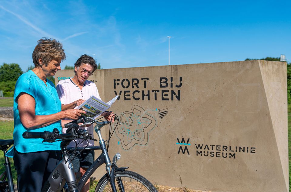 Een man en een vrouw staan met hun fietsen bij een grijs bord waarop 'Fort bij Vechten' en 'Waterliniemuseum' staat. Ze bekijken samen een folder.