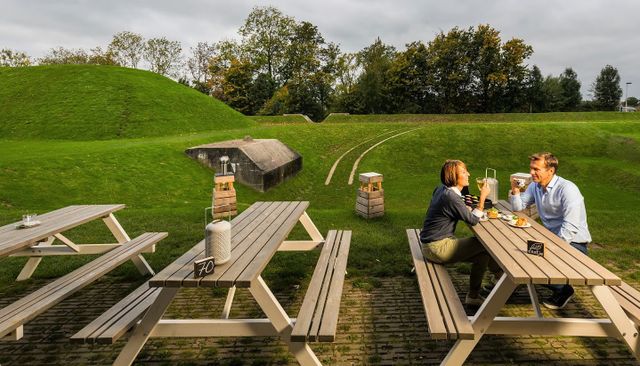 Een man en een vrouw zitten op een picknickbank in het gras te genieten van eten en drinken. op de achtergrond zie je onderdelen van het fort.