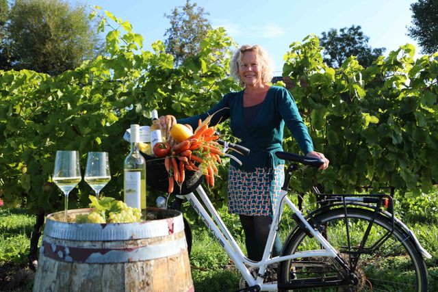 een mevrouw en een fiets met daarop een mandje vol eten en een fles wijn
op de voorgrond een houten ton met daarop twee wijnglazen