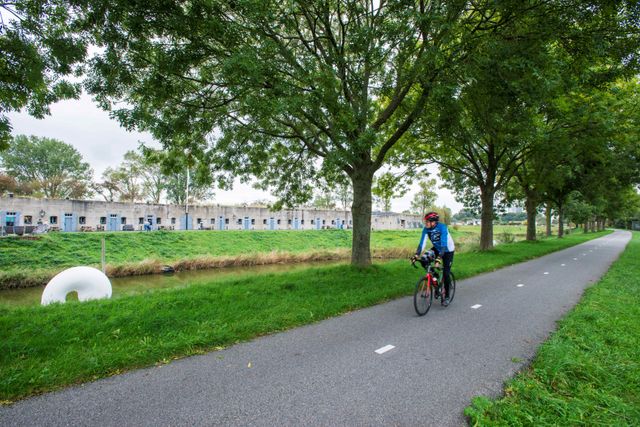 Een wielrenner op een asfalt fietspad langs een rij bomen en een waterpartij. Aan de andere kant van het water is een betonnen fort te zien. In het water ligt een wit kunstwerk.
