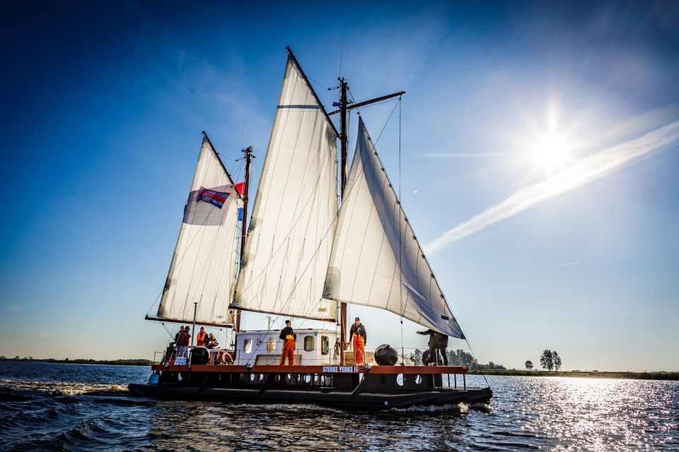Vlot Sterke Yerke IV vaart op het Friese water met de zon schijnend door de zeilen en de bemanning aan dek.