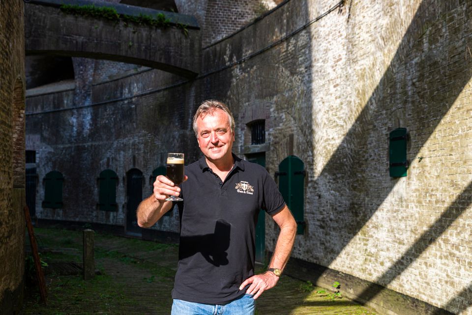 Een man met een zwart shirt staat in de omgang van een fort met een glas bier in zijn hand.