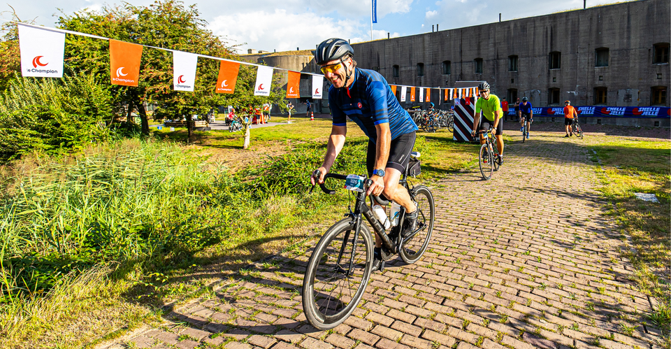 Verschillende personen op wielrenfietsen rijden weg over een klinkerpaadje met op de achtergrond een betonnen fort van de Stelling van Amsterdam. Langs het pad hangen witte en oranje vlaggetjes met daarop de tekst 'Le Champion'.