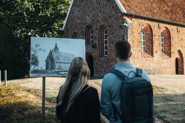 Een man en een vrouw kijken naar de schets van het kerkje van Zweeloo dat vlak voor dit kerkje op een bord is geplaatst.