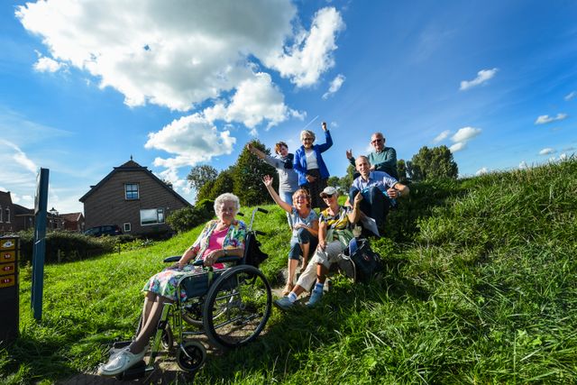 Foto van een familie die gezellig met oma in een rolstoel aan het wandelen zijn in de natuur.