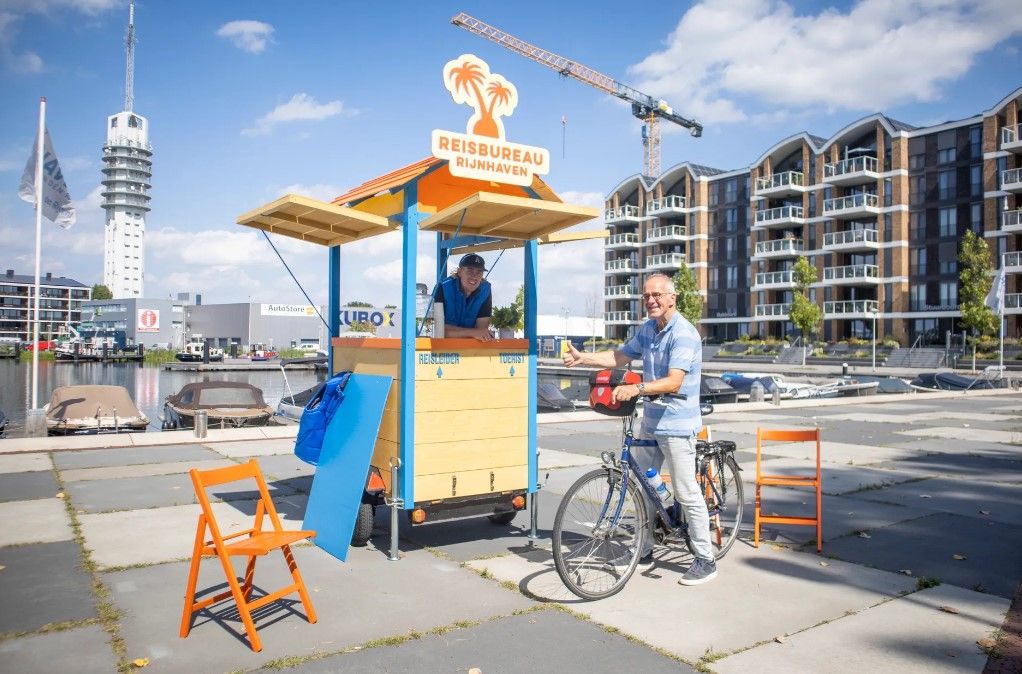 Een klein kraampje met de tekst reisbureau Rijnhaven en met daarnaast een man op zijn fiets die lacht en zijn duim opsteekt.