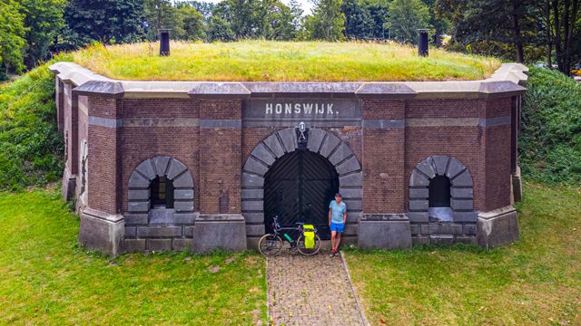 Een fietser staat met zijn fiets voor het poortgebouw van een fort. Boven de deur staat: Fort Honswijk.