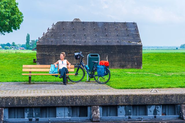 Een vrouw zit op een houten bankje. Tegen het bankje staat haar fiets geleund. Achter haar is een betonnen groepsschuilplaats (bunker) te zien.