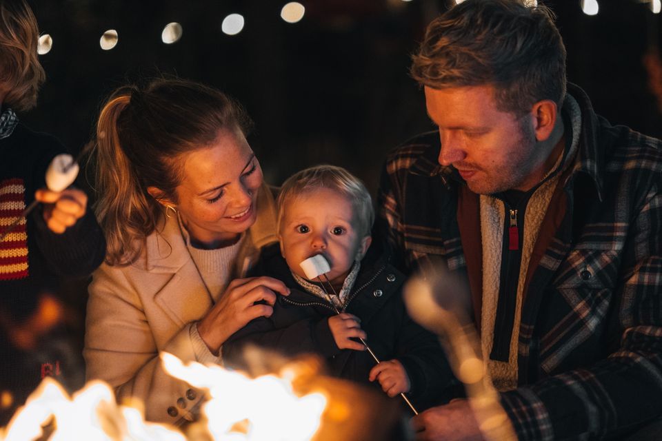 Vader en moeder zitten met een kindje dat van een marshmallow eet bij het kampvuur.