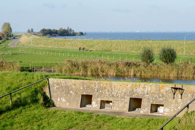 Een betonnen onderdeel van een fort vlakbij een dijk met daarachter een meer. Langs de dijk rijden twee fietsers.
