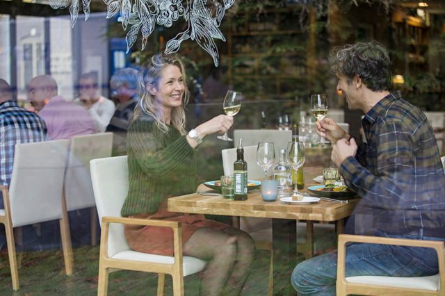 Een man en vrouw zitten aan tafel te dineren en proosten met een glas wijn.