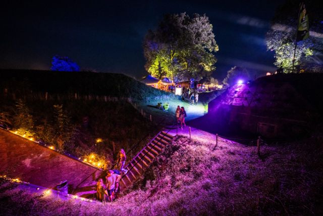 Een donkere foto waarop je mensen ziet lopen over een fort dat is uitgelicht in mooie kleuren als paars, groen en blauw.