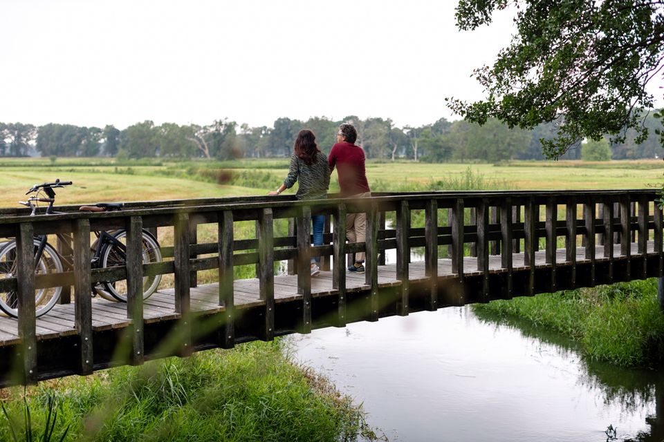 Een man en vrouw hebben hun fietstocht gepauzeerd en genieten vanaf een brug van het uitzicht op de natuur.