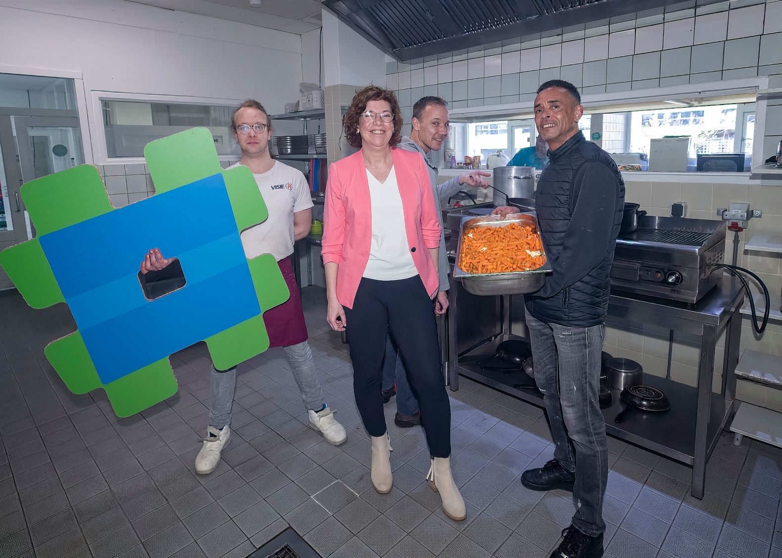 Een vrouw met drie mannen in de keuken waarvan een man een bak eten vasthoud dan houd er meneer een pan vast en nog een jongen ober met het HierisALPHEN logo.
