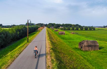 Een vrouw fietst over een asfalt fietspad op een dijk, naast de dijk liggen verschillende betonnen groepsschuilplaatsen uit de Nieuwe Hollandse Waterlinie.
