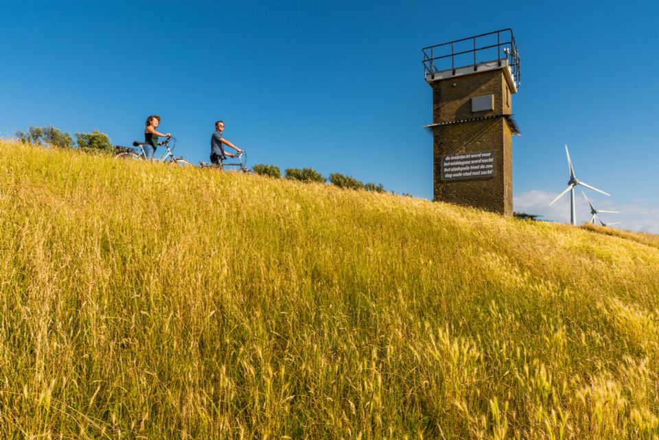 Een dijk waar een kleine toren op staat, twee fietsers pauzeren om het uitzicht te bewonderen, op de achtergrond staan een paar windmolens. In de Noordoostpolder.