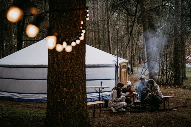 Een aantal mensen zitten bij het kampvuur naast een yurt in het bos.