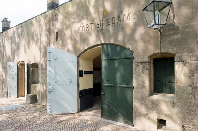 Het betonnen fort bij Edam. De metalen buitendeuren staan open en boven de deur staat 'Fort bij Edam' uitgehouwen in steen. Naast de deur hangt een ouderwetse lantaarn.