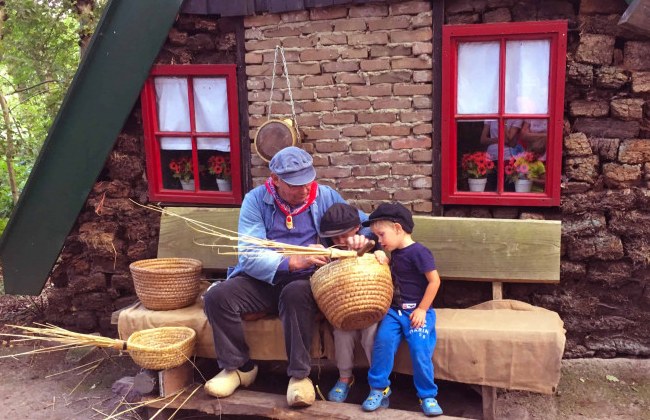 Een mandenmaker in openluchtmuseum Veenpark laat 2 jongentjes zien hoe je manden vlecht met riet. Ze zitten op een bankje voor een echte Drentse plaggenhut.