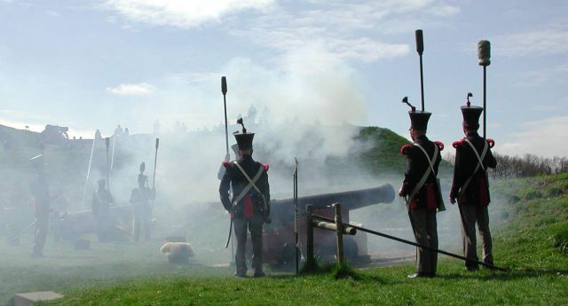 Een aantal mannen in ouderwetse uniforms staan in kruitdampen terwijl zij kanonnen afschieten in een fort.