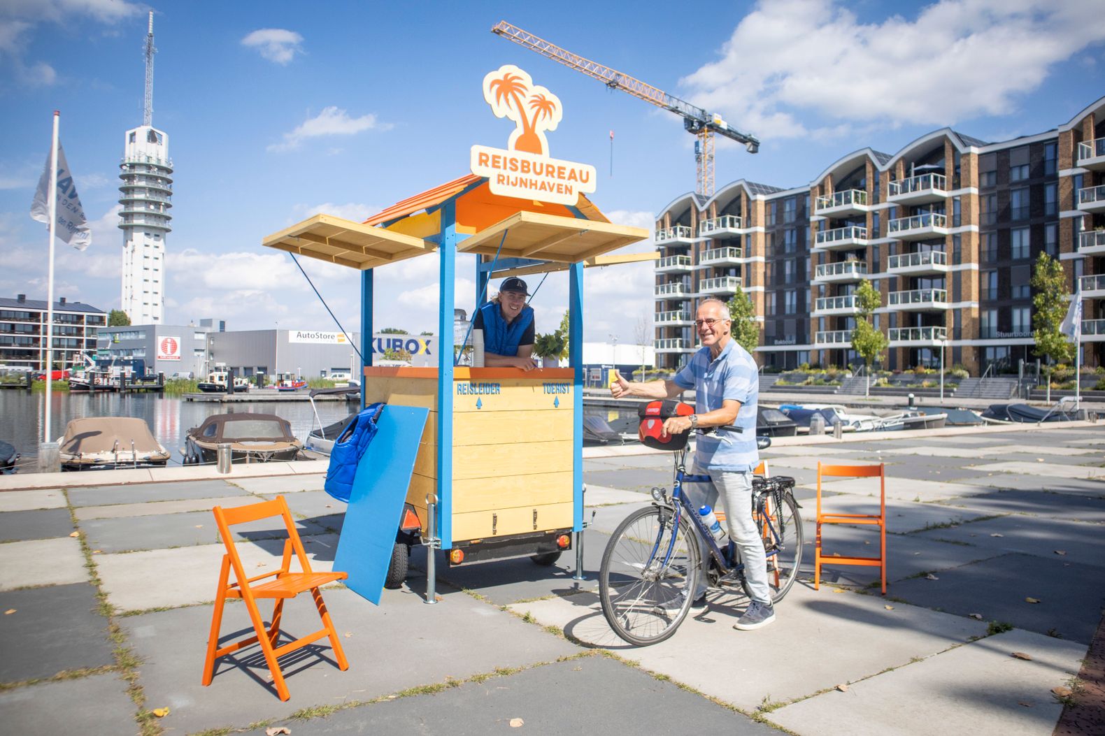 Een vrouw achter het reisbureau kraampje van Rijnhaven met een meneer op zijn fiets die daarnaast staat.