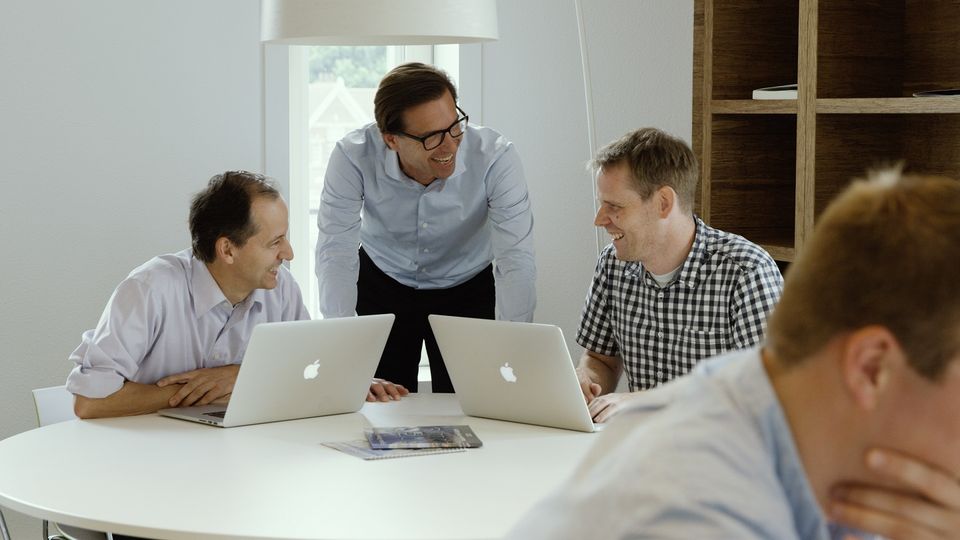 Foto van drie mannen op kantoor achter een laptop die naar elkaar lachen.