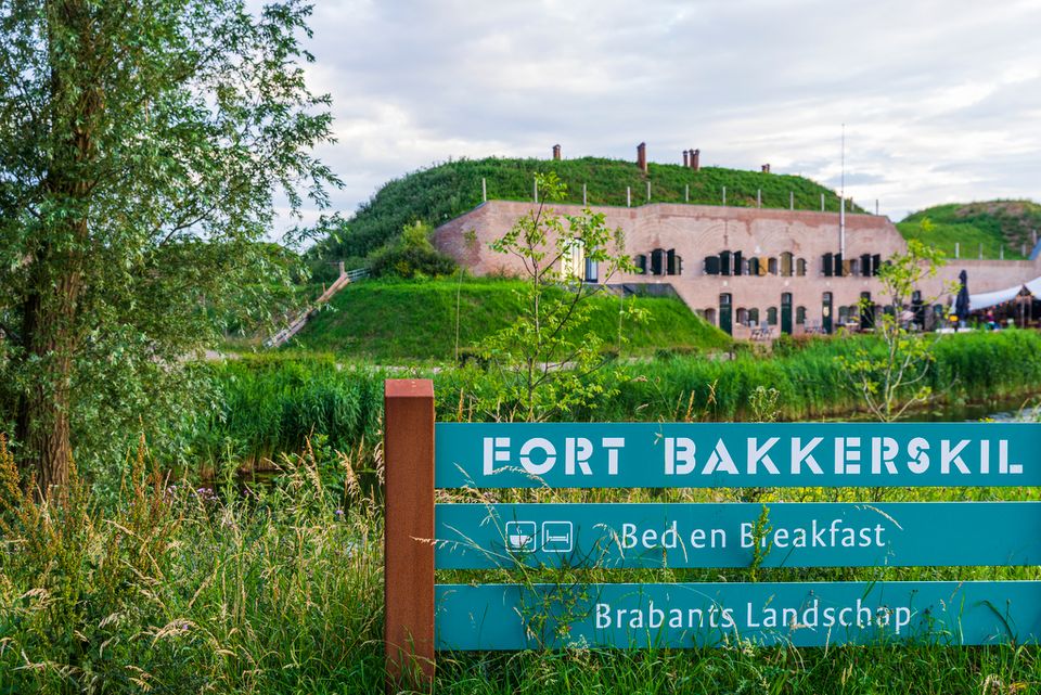 Een groot bakstenen fort gefotografeerd van de voorkant. In het gras staat een groot blauw bord met witte letters: Fort Bakkerskil, Bed & Breakfast, Brabants Landschap.
