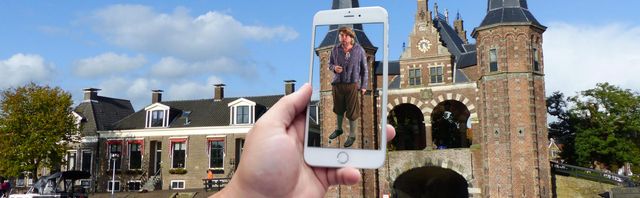 Op de telefoon is de app Mear Fryslân te zien. De telefoon wijst naar de Waterpoort in Sneek, en op het beeld zie je een oudere meneer in klederdracht staan.