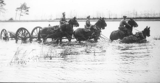 Drie soldaten leiden vier paarden en een kanon door een onder water gelopen gebied.