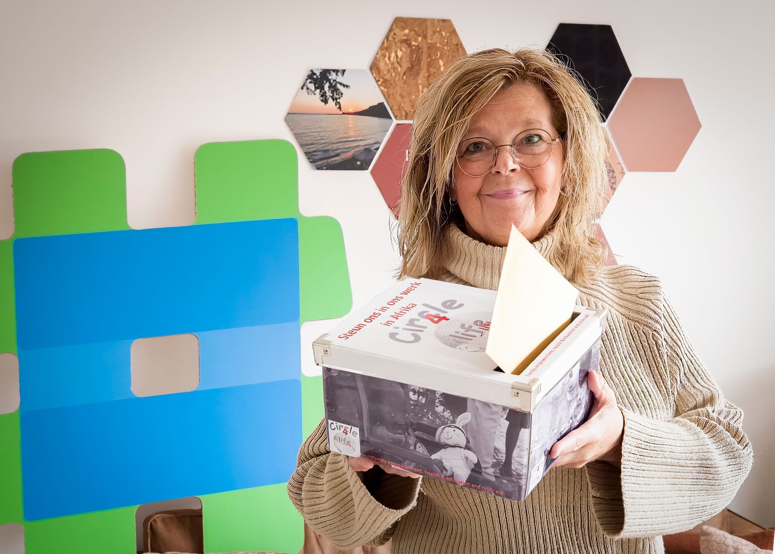 Een vrouw van de organisatie Circle of Life doneer box vast houd met op de achtergrond het logo van HierisALPHEN.