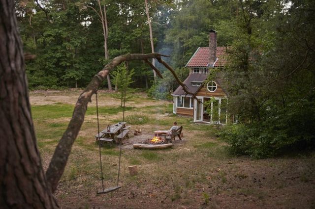 Een vrouw zit bij een kampvuur naast een vakantiehuisje in het bos.