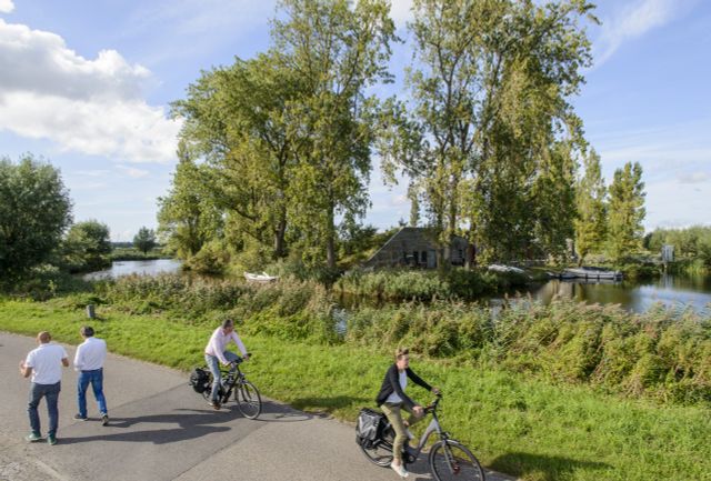 Twee fietsers en twee wandelaars lopen en fietsen over een asfalt pad aan het water. In het water is een betonnen fort omgeven door bomen zichtbaar.