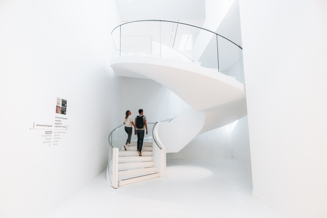 Een man en een vrouw beklimmen de witte wenteltrap in het Drents Museum in een ruimte met witte muren, een witte vloer en een wit plafond.
