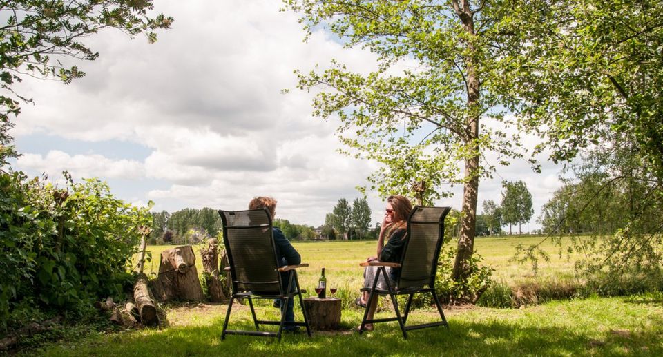 Een man en een vrouw zitten samen op kampeerstoelen, tussen hen in een houtblok dat als tafel dient met daarop een fles wijn en twee glazen. Ze kijken uit over de uitgestrekte natuur in de omgeving.