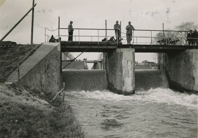 een zwart-witfoto waarop het gebruik van een inundatiesluis zichtbaar is. Je ziet soldaten bovenop de sluis staan die open is, waardoor er water doorheen stroomt.