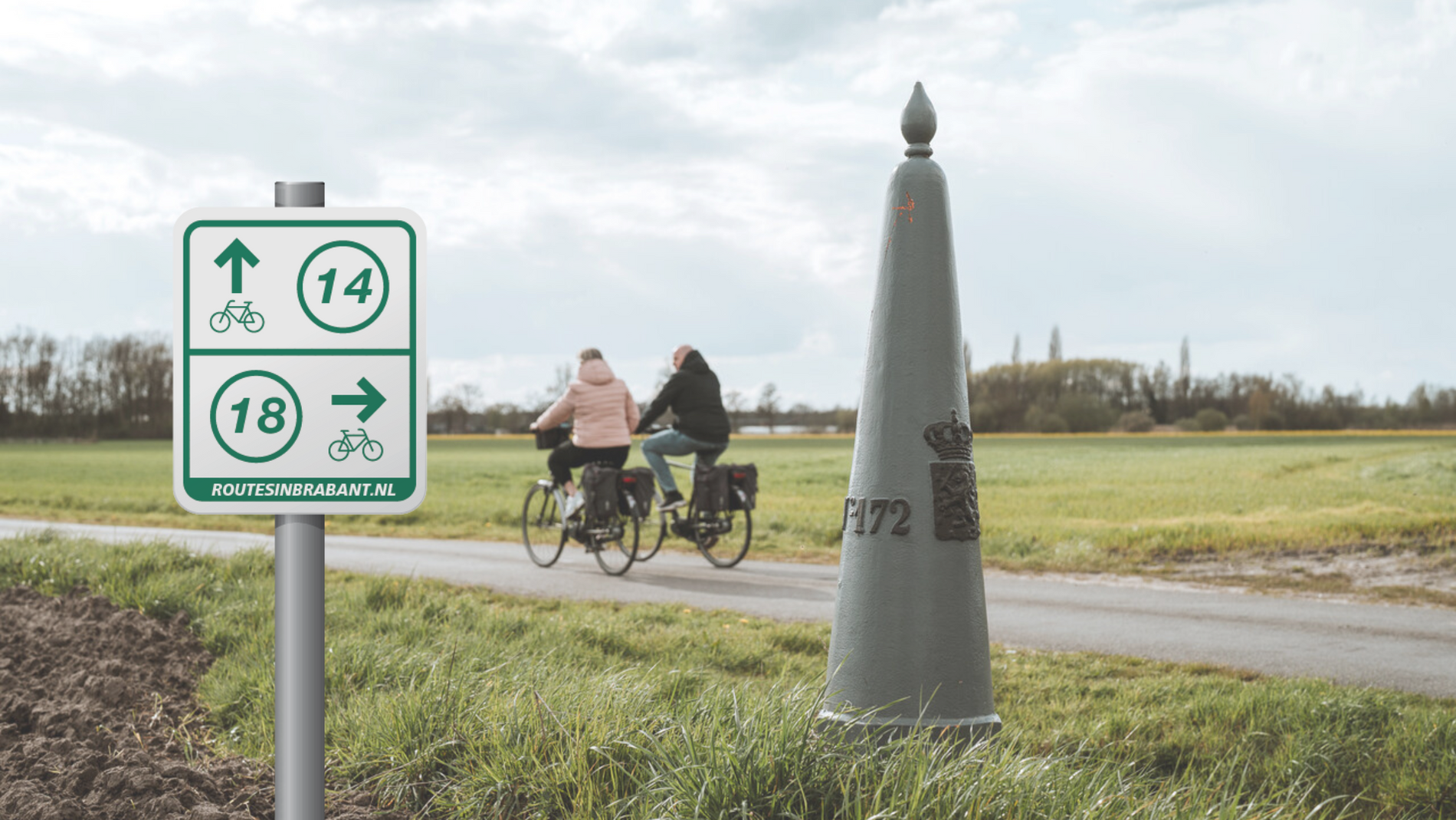 Routepaal naast grenspaal met fietsende mensen op de achtergrond