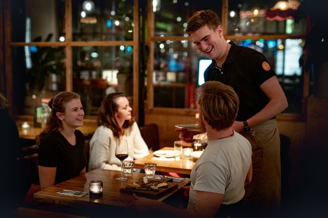 De serveerder van het restaurant staat aan tafel bij twee bezoekers. Ze lachen met elkaar.