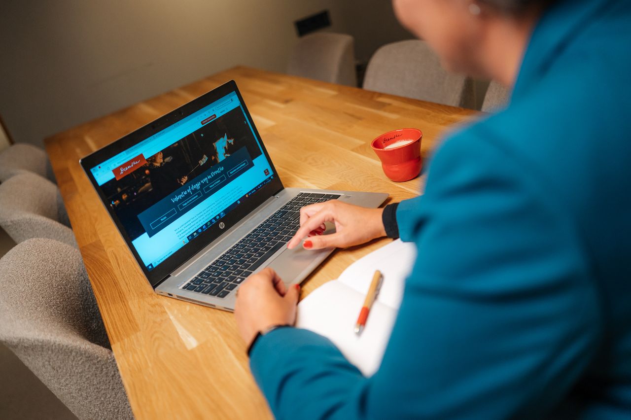 Een vrouw zit achter de laptop aan de keukentafel met een kopje koffie ernaast.
