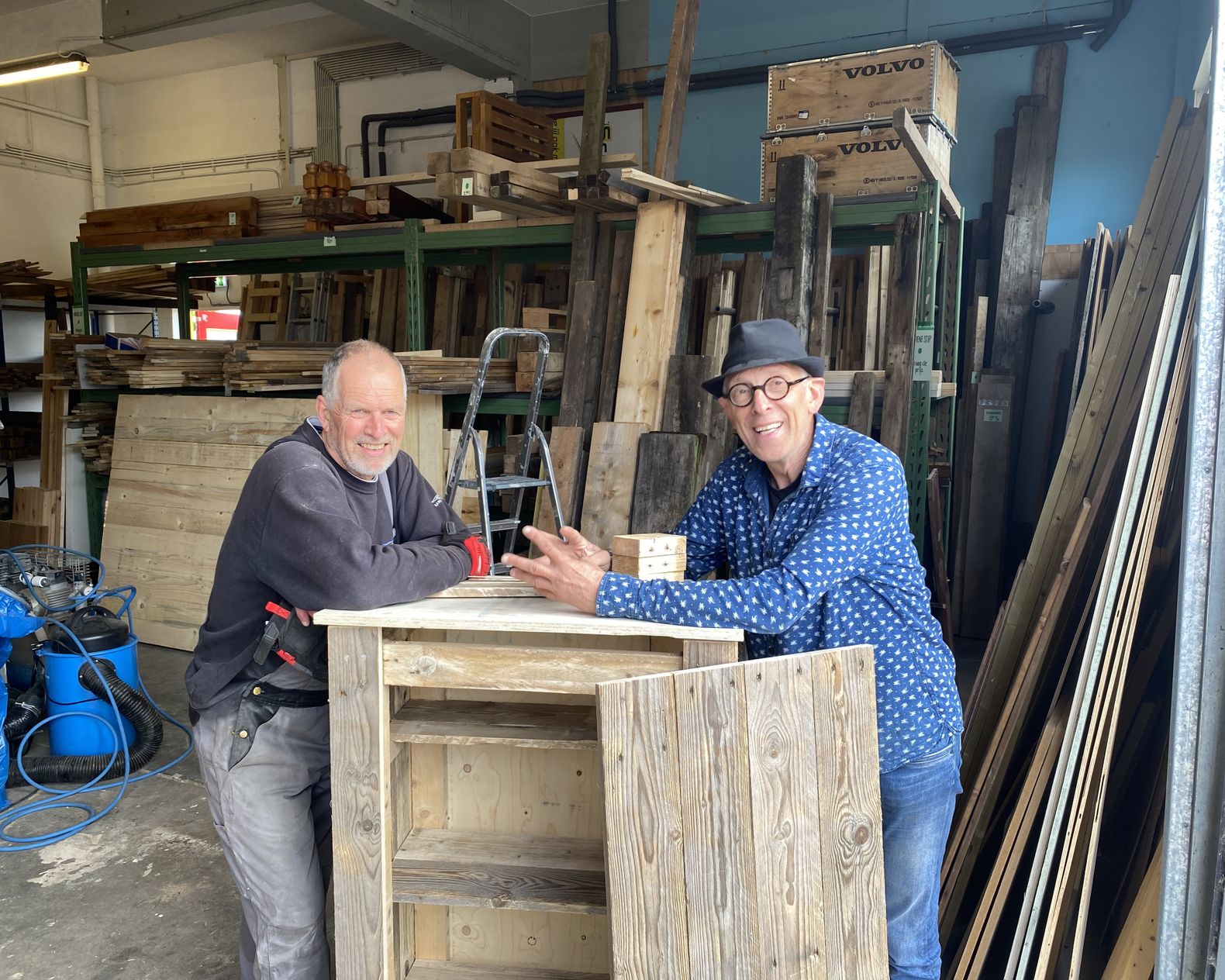 Twee mannen die staan bij een hout meubelstuk omringt door allemaal verschillende houtplanken.